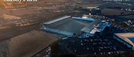 An aerial view of Exeter Sandy Park, featuring a large stadium, parking area, and surrounding landscape.