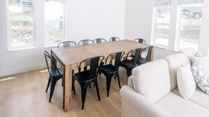 Une pièce spacieuse avec une table à manger en bois entourée de chaises, un canapé blanc et de grandes fenêtres donnant sur l’extérieur.
