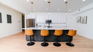 Une cuisine moderne dotée d’un îlot central, de quatre tabourets de bar, de meubles blancs, d’un réfrigérateur en acier inoxydable et de luminaires suspendus.