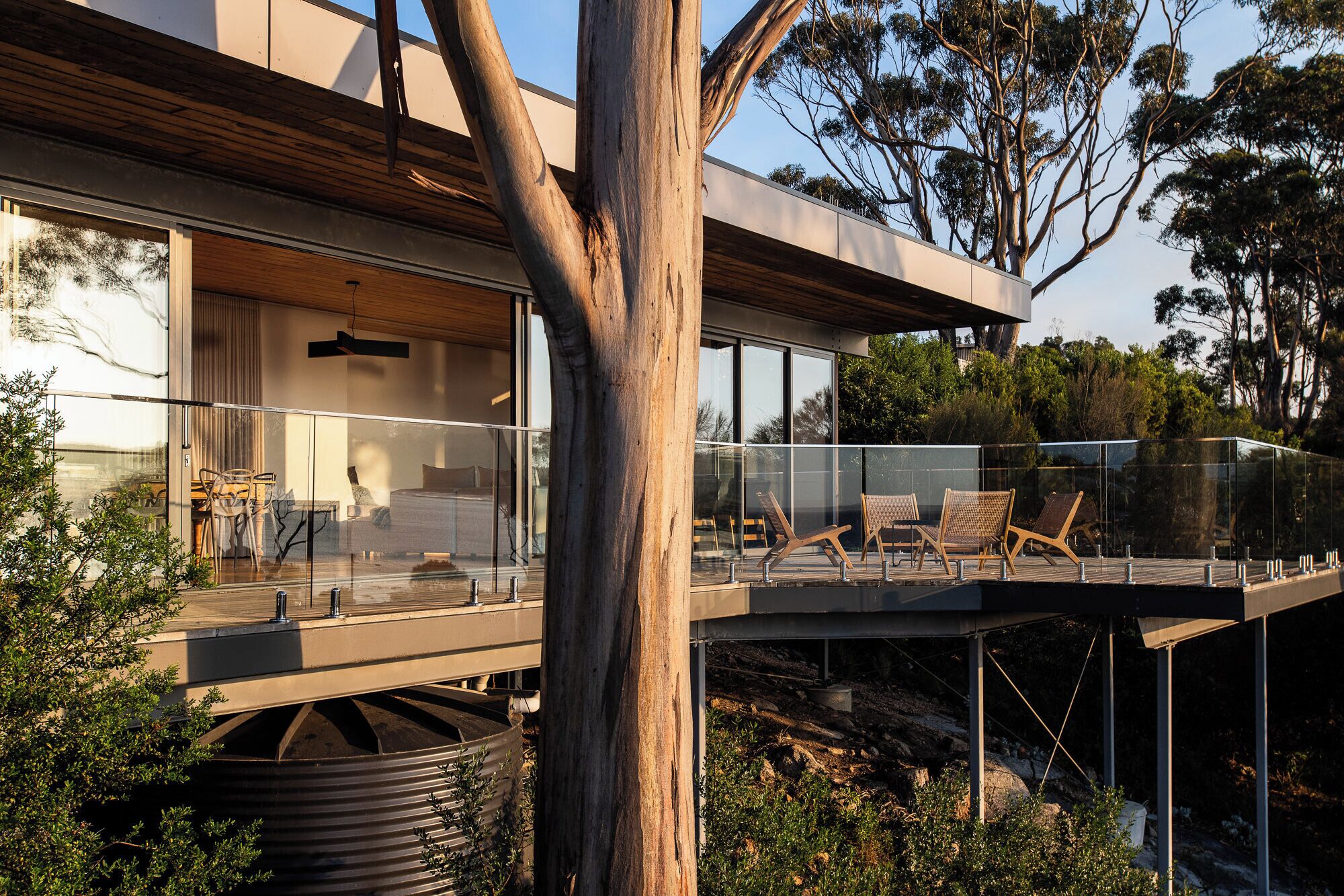 A modern house with a glass balcony, wooden deck, and a view of the surrounding trees.