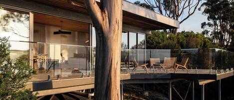 A modern house with a glass balcony, wooden deck, and a view of the surrounding trees.