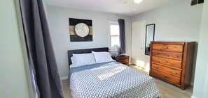 Une chambre à coucher comprenant un lit, une commode, une table de chevet, une fenêtre avec des rideaux et une œuvre d’art accrochée au mur.