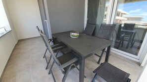 En balkon med et mørkegråt bord og fire stole, en lille glasskål med grøntsager, og udsigt til bygninger gennem glasdøren.