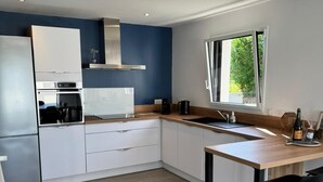 A modern kitchen with white cabinets, a stainless steel range hood, a built-in oven, a coffee maker, and a window providing natural light. - The iodine parenthesis. House with garden - Near sea (Plozévet)