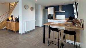 A modern kitchen with a built-in oven, a dining area with a wooden table and bar stools, and a staircase leading to another level. - The iodine parenthesis. House with garden - Near sea (Plozévet)