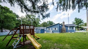 Un jardin avec une structure de jeu en bois, une cheminée en pierre et une maison bleue.
