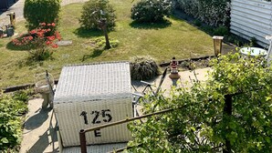 Un jardín residencial con una camioneta blanca estacionada en la entrada, un edificio blanco con el número 125 a la vista, y una variedad de follaje, incluyendo arbustos y plantas con flores.
