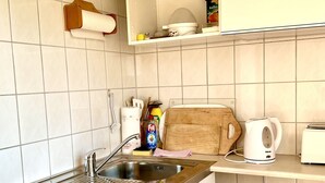 Cocina compacta con lavabo, estufa y un refrigerador pequeño. Hay repisas sobre el lavabo con distintos utensilios de cocina.