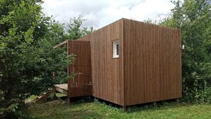 A small, wooden cabin with a single window and a small door, surrounded by greenery.