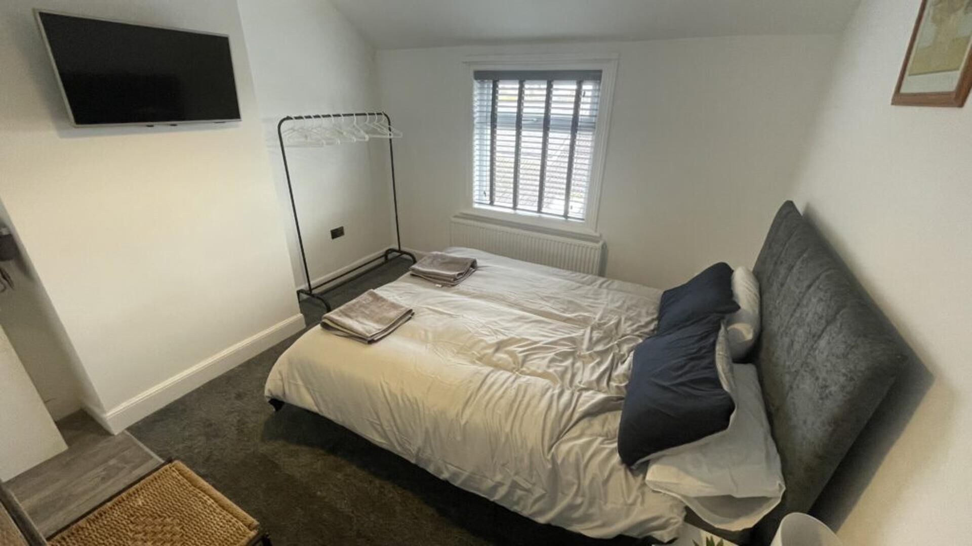 A bedroom with a bed, a TV mounted on the wall, a window with blinds, and a radiator.