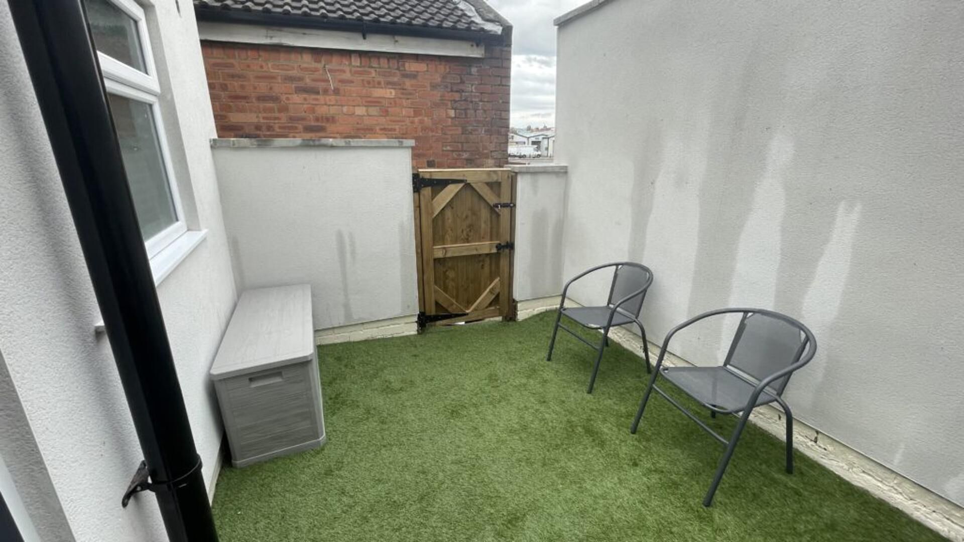A small enclosed outdoor area with artificial grass, two metal chairs, a wooden gate, and a white wall.