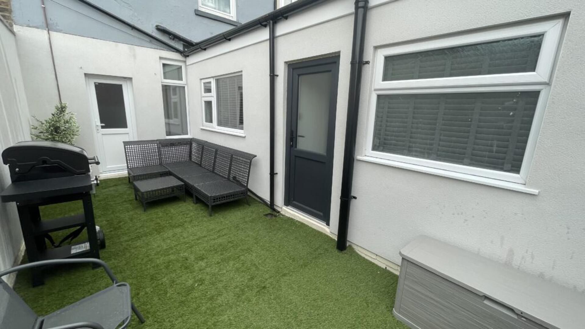A small backyard with artificial grass, a black grill, a black outdoor sofa, and a door.