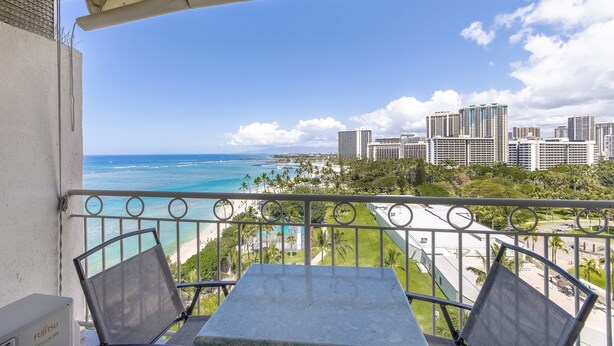 A balcony with a table and chairs, overlooking a beach and cityscape. - Oceanfront Studio | Spectacular Views | Full Kitchen | Nearby Parking(Fee) (Honolulu)
