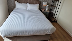 Ein ordentlich bezogenes Bett mit einem gestreiften Überwurf, ein Nachttisch mit einer Lampe und einer Fernbedienung sowie ein Holzboden.
