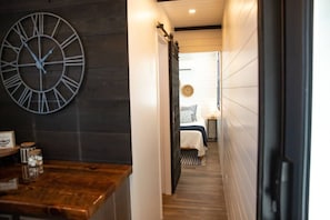 A hallway leading to a bedroom with a bed, a bedside table, and a wall clock.