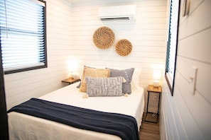 A bedroom with a bed, bedside tables, wall-mounted air conditioning, and decorative woven wall art.