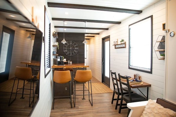 A modern kitchen with a large clock, wooden bar stools, and a dining area with chairs.