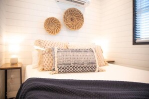 Ein ordentlich eingerichtetes Schlafzimmer mit einem Bett, Kissen, einem Nachttisch mit Lampe und einer Wand-Klimaanlage.