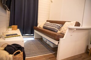 A compact living space with a sofa, a small table with books, a basket, and a window with curtains. - New! Endless Sunsets-Container Home (Alpine)
