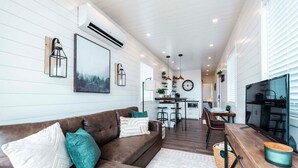 A modern living room with a brown leather sofa, a dining area, a kitchen, and a TV. - The Stardust Stunning Container Home (Alpine)