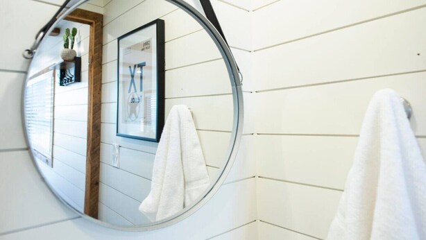 Une salle de bain avec un miroir rond, des murs recouverts de carreaux blancs et un tableau encadré.