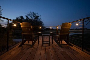 A deck with two wooden chairs and a table, surrounded by a railing with string lights. - The Lone Star Shipping Container | Rooftop Patio (Fredericksburg)