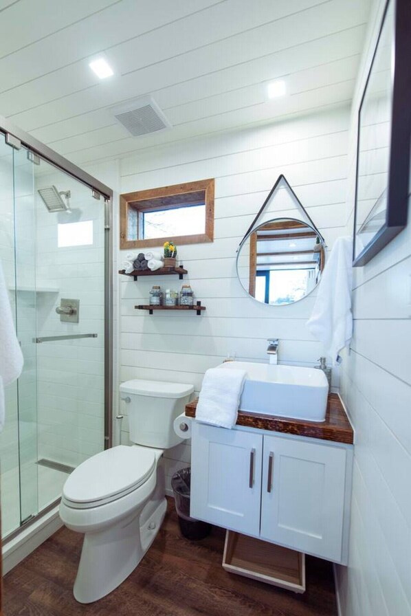 A bathroom with a glass shower enclosure, a toilet, a sink with a mirror, and a wooden vanity. - The Lone Star Shipping Container | Rooftop Patio (Fredericksburg)