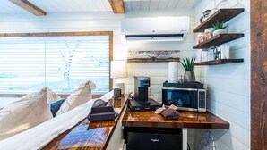 A compact kitchen with a microwave, coffee maker, and a refrigerator. There is a window with blinds, a wooden ceiling, and a shelf with kitchen items. - The Lone Star Shipping Container | Rooftop Patio (Fredericksburg)