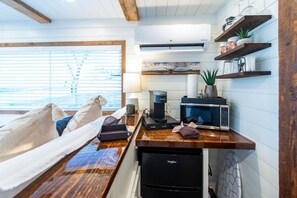 A compact kitchen with a microwave, coffee maker, and a refrigerator. There is a window with blinds, a wooden ceiling, and a shelf with kitchen items. - The Lone Star Shipping Container | Rooftop Patio (Fredericksburg)