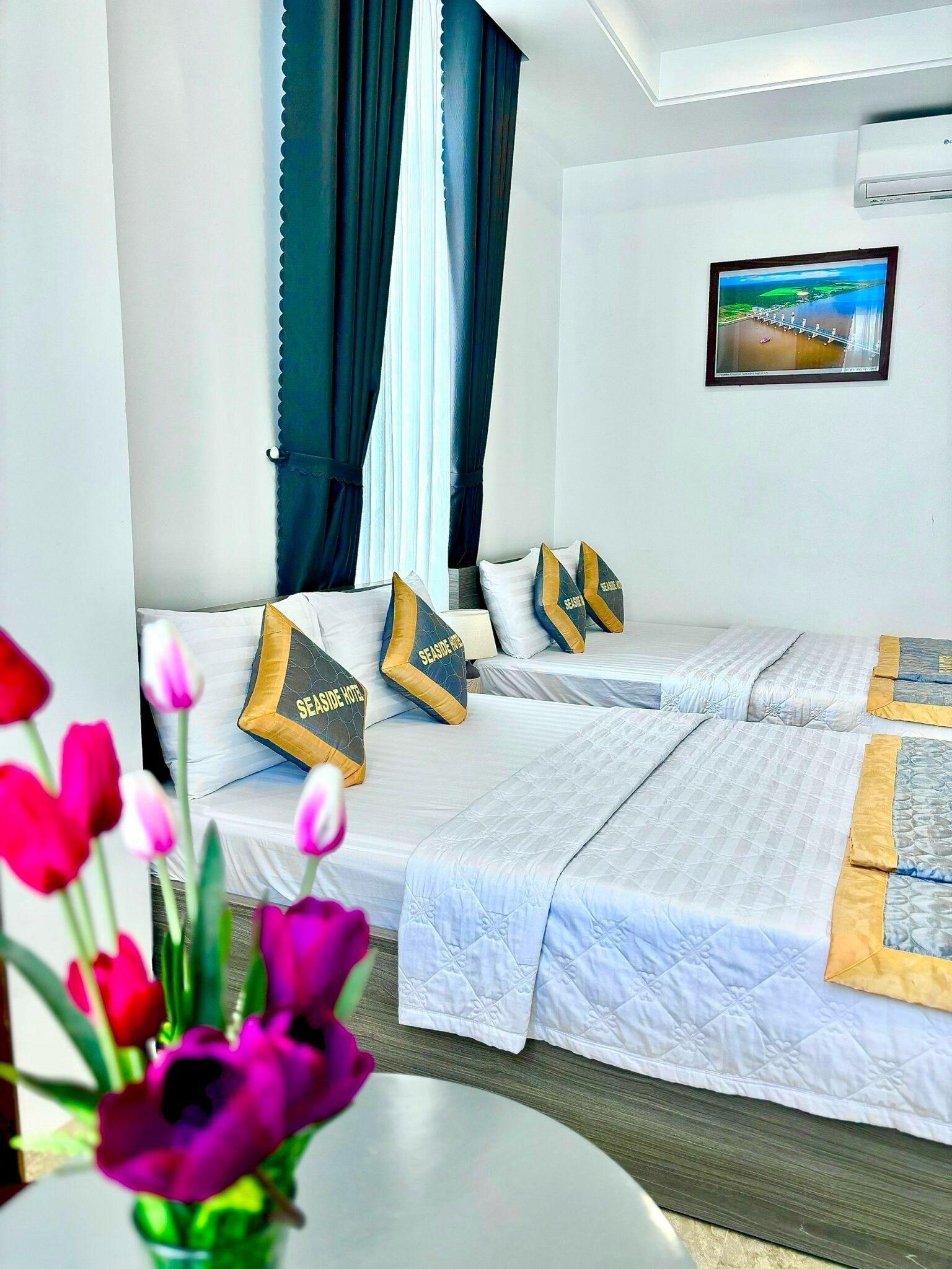 A hotel room with two beds, a large window with curtains, a painting on the wall, and a vase of flowers on a table.