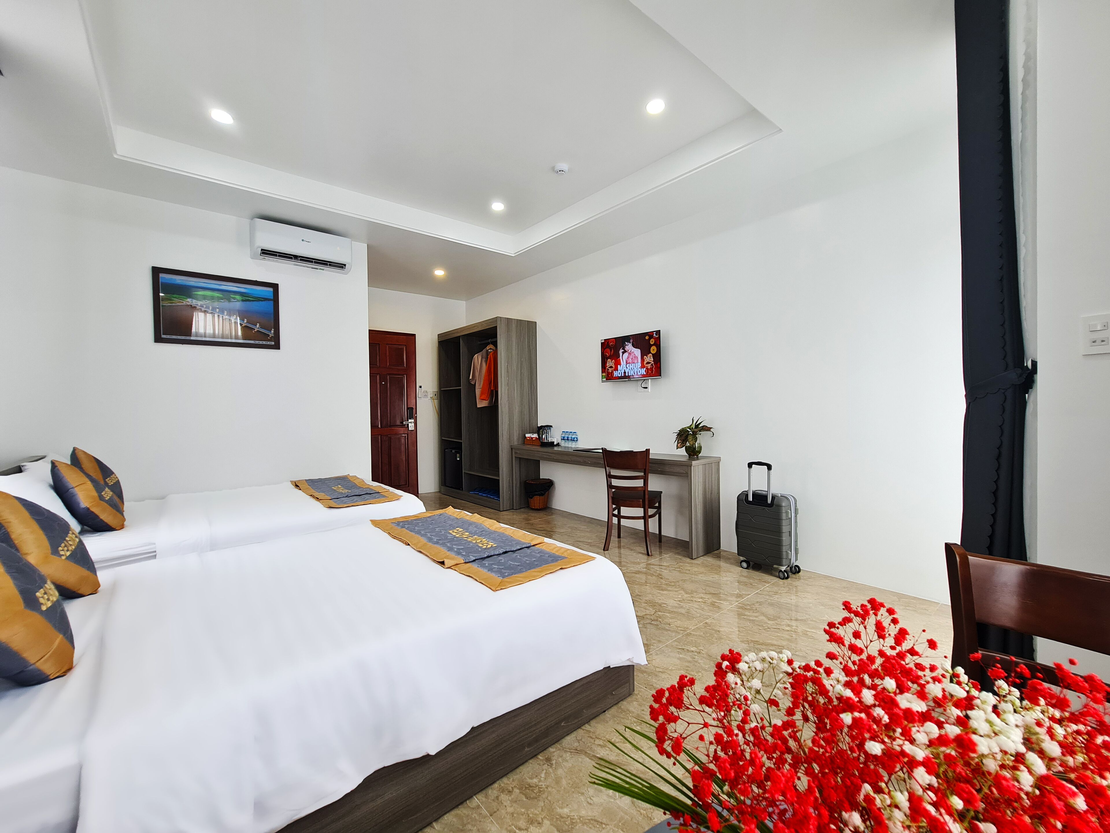 A hotel room with two beds, a desk, a chair, a TV, and a suitcase.