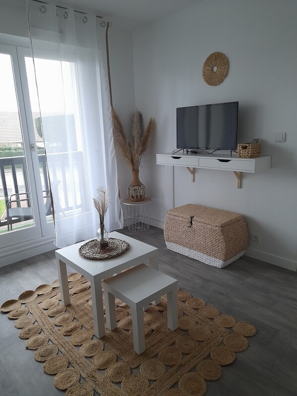 A modern living room with a white TV stand, a woven pouf, a small table with a vase, and a large woven rug.