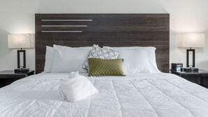 Un lit bien fait, avec du linge de lit blanc, une couette matelassée et une serviette blanche posée sur le lit. Il y a deux tables de chevet avec des lampes et un coussin décoratif sur le lit.