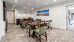 Un vaste espace dédié à la salle à manger, avec des tables et des chaises en bois, une cuisine à l’arrière-plan et une grande fenêtre donnant sur un immeuble.