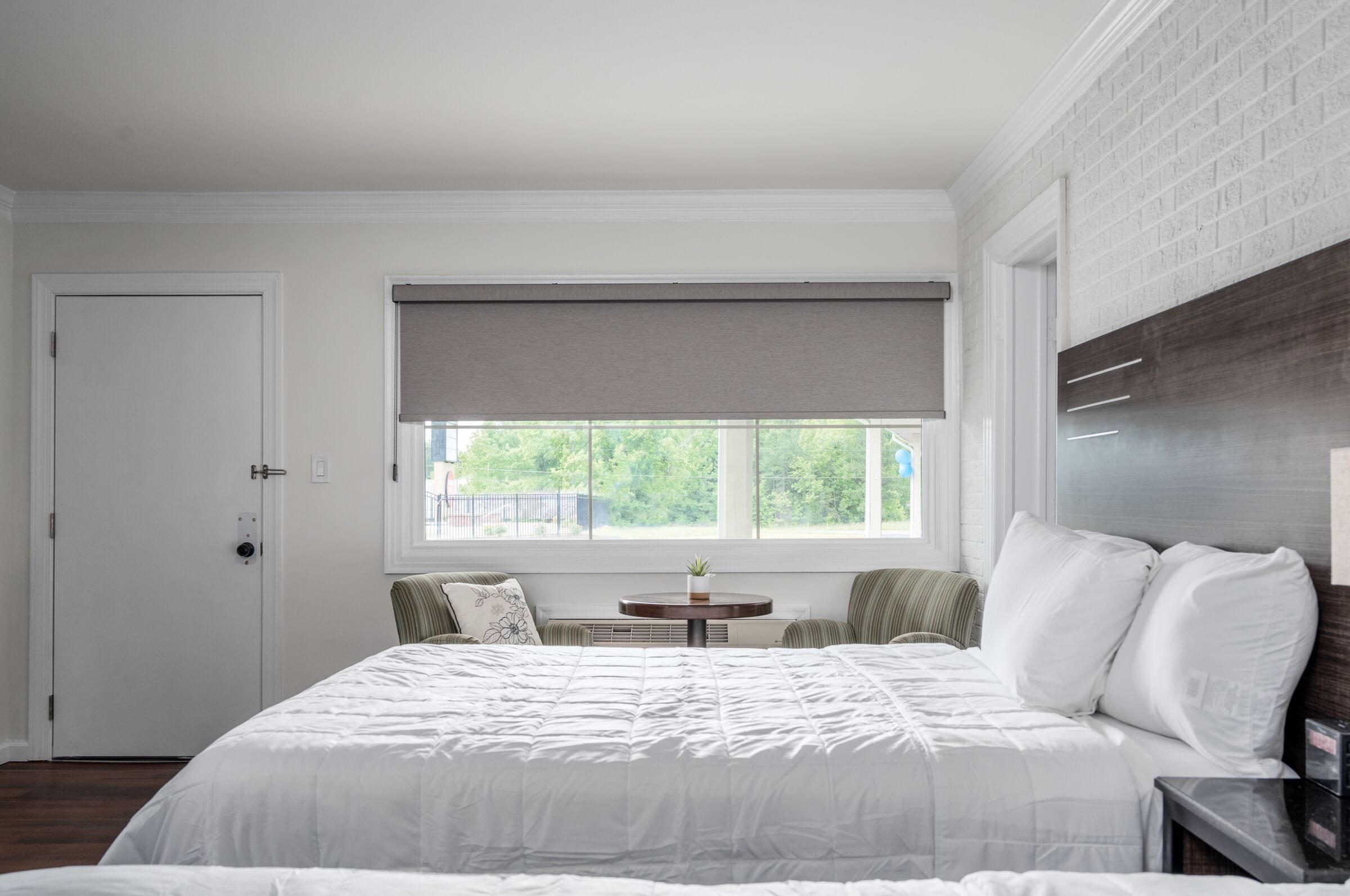 A bedroom with a large bed, a window with a view of greenery, a small table with a plant, and a door.