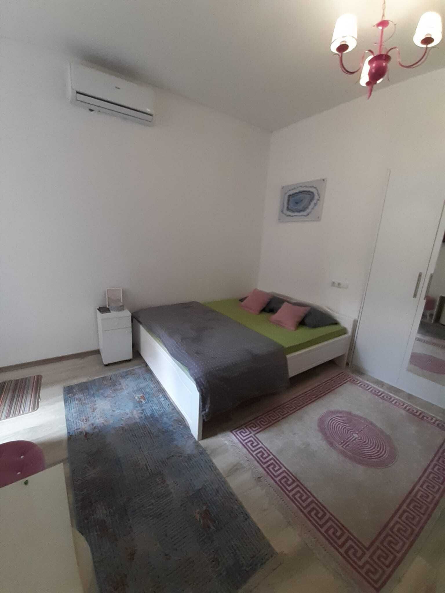 Quarto pequeno e simples com uma cama, mesa de cabeceira, ar condicionado, tapete e luminária de teto.