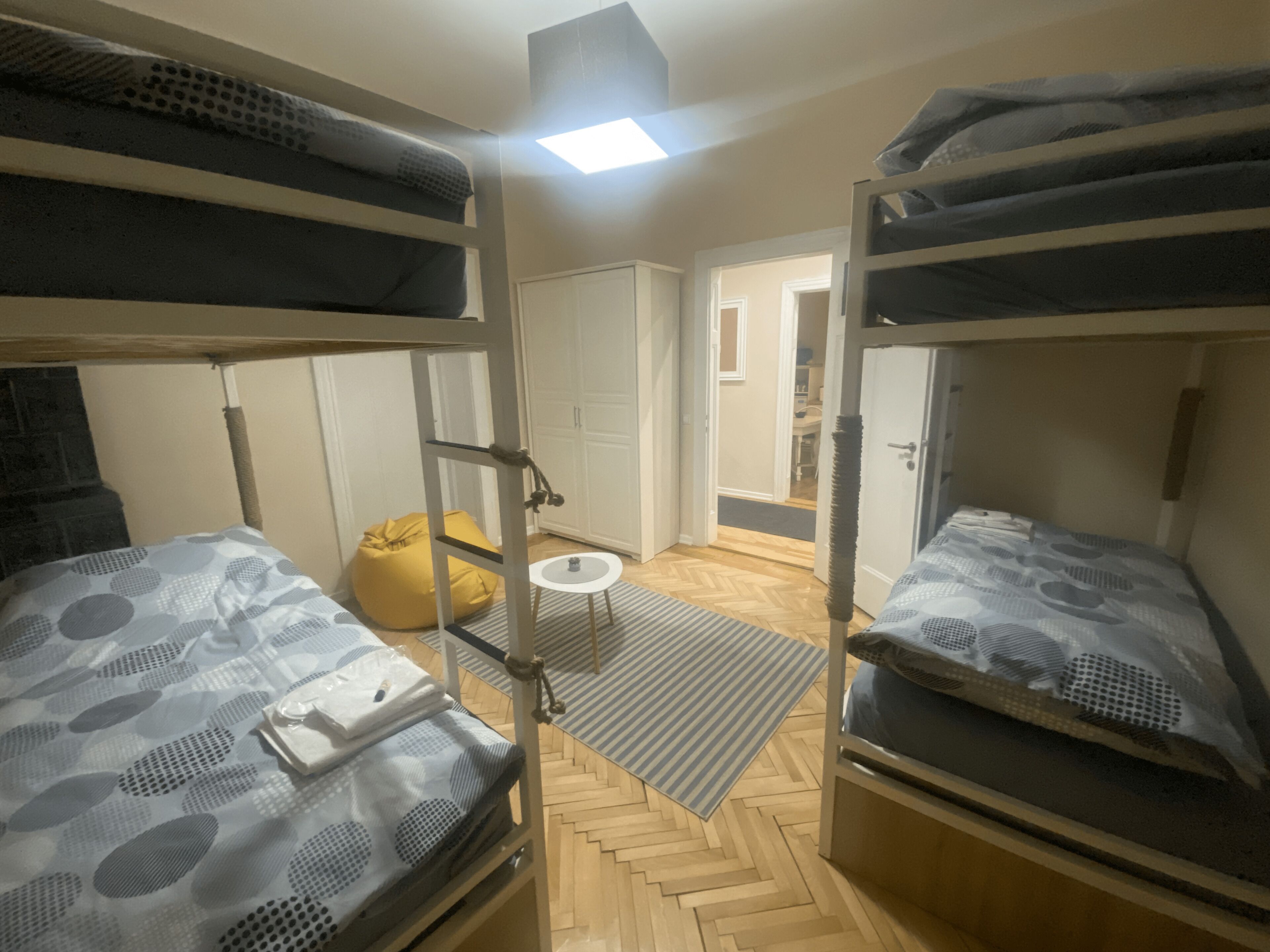 A dormitory room with bunk beds, a small round table, a yellow bean bag, and a door leading to another room.