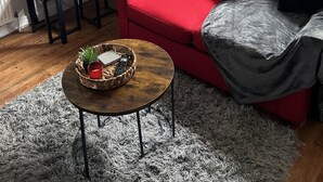 A modern living room with a red sofa, a small wooden table with a basket, a grey furry rug, and a large window with curtains. - Inviting 1-bed Unit in Watford (Watford)