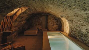 Relax in a serene spa setting with a rectangular pool, wooden bench, and stone bench with cushioned seat, surrounded by an arched stone ceiling and walls.