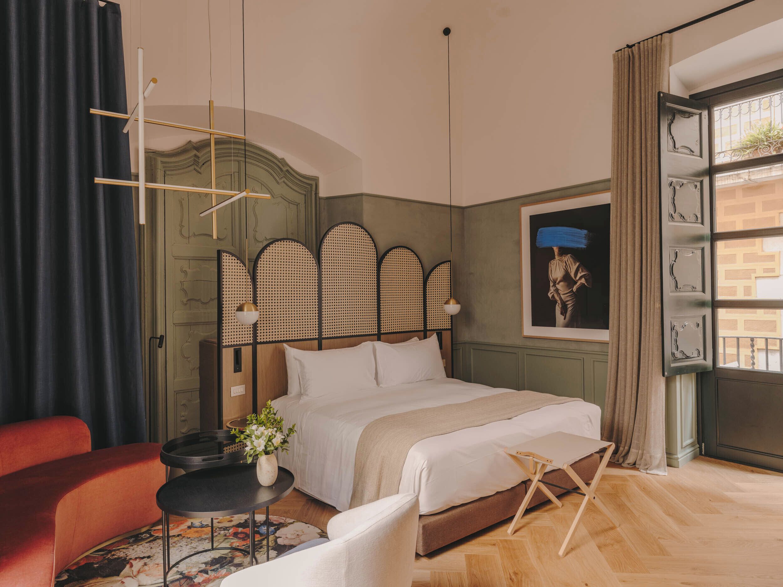 Un dormitorio con una cama, una mesita con un florero, una silla roja y una ventana con cortinas.