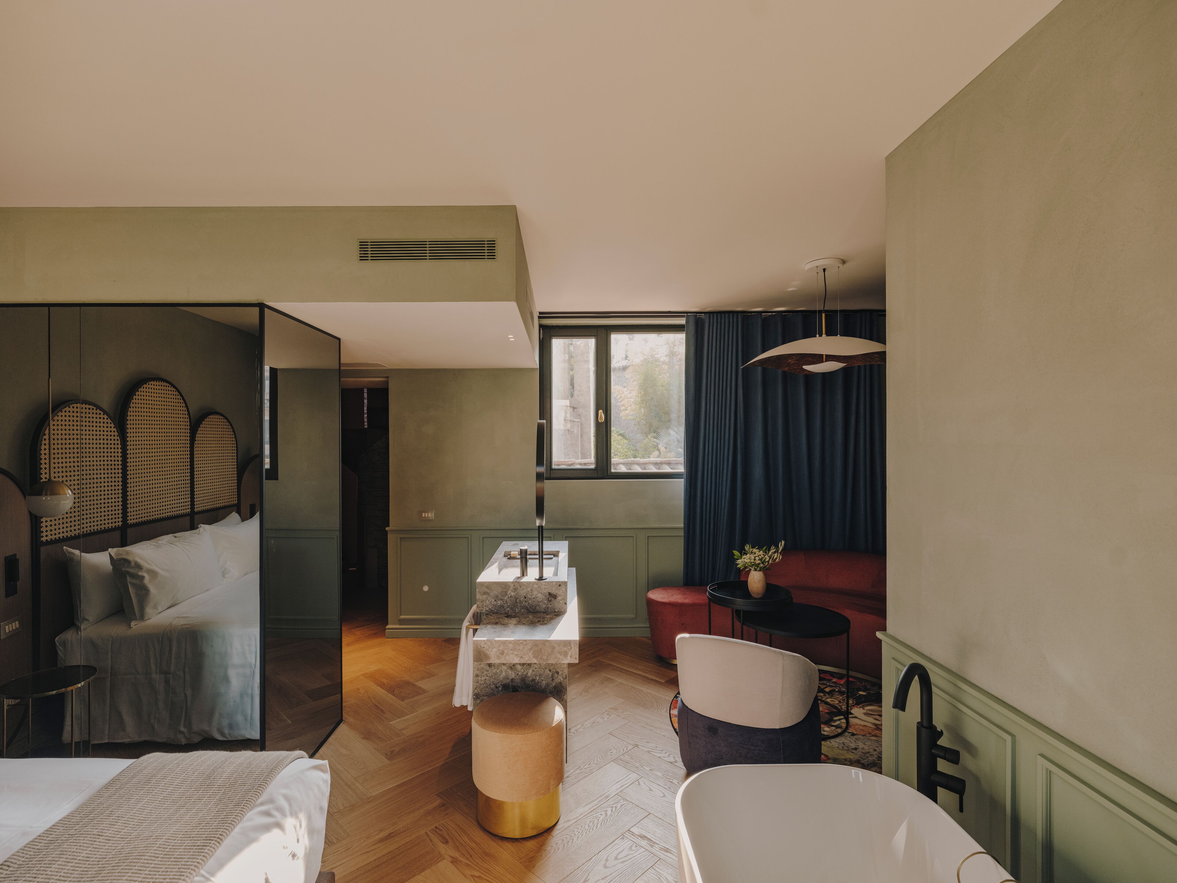 A modern hotel room with a large bed, a sitting area with a sofa and a small table, a bathroom with a bathtub, and a large window providing natural light.