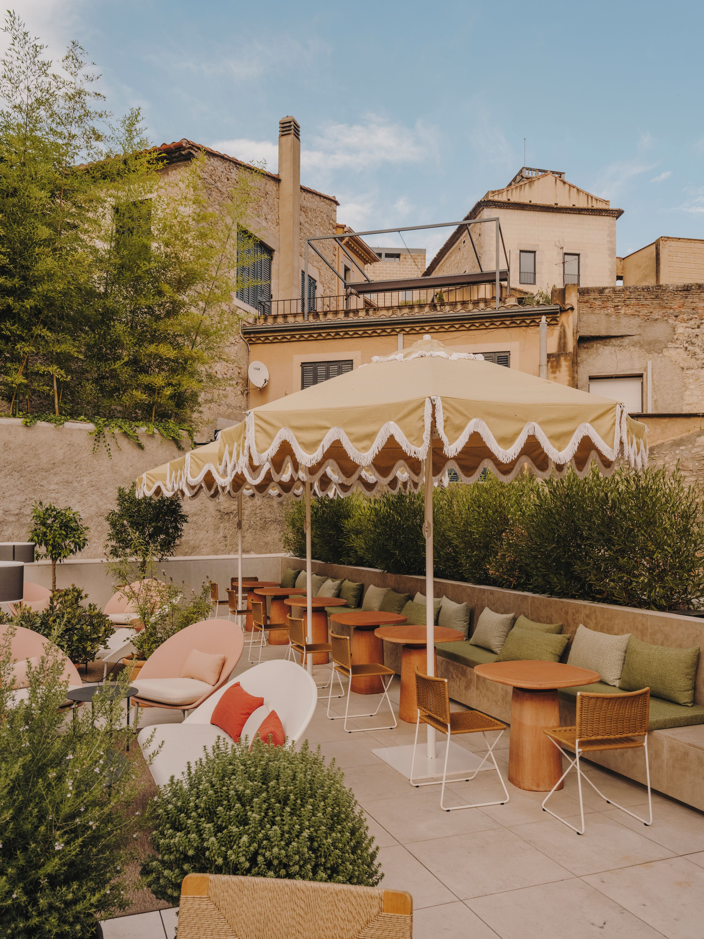 An outdoor seating area with round tables and chairs, surrounded by greenery and a building in the background.