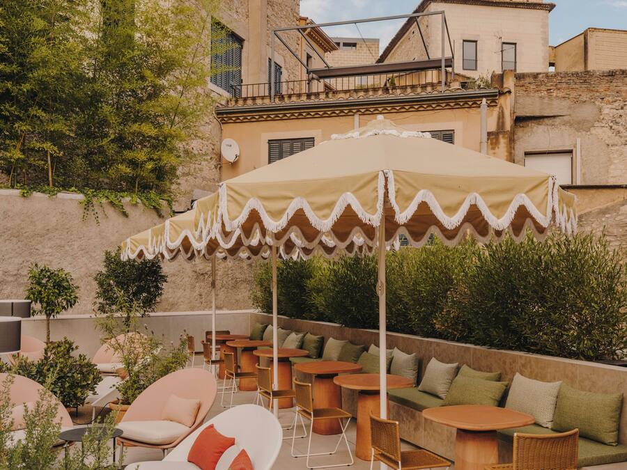 An outdoor seating area with round tables and chairs, surrounded by greenery and a building in the background.