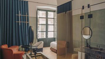 Quarto de hotel moderno com parede de destaque em pedra, box de chuveiro de vidro, área de estar com uma mesinha e uma janela grande com cortinas.