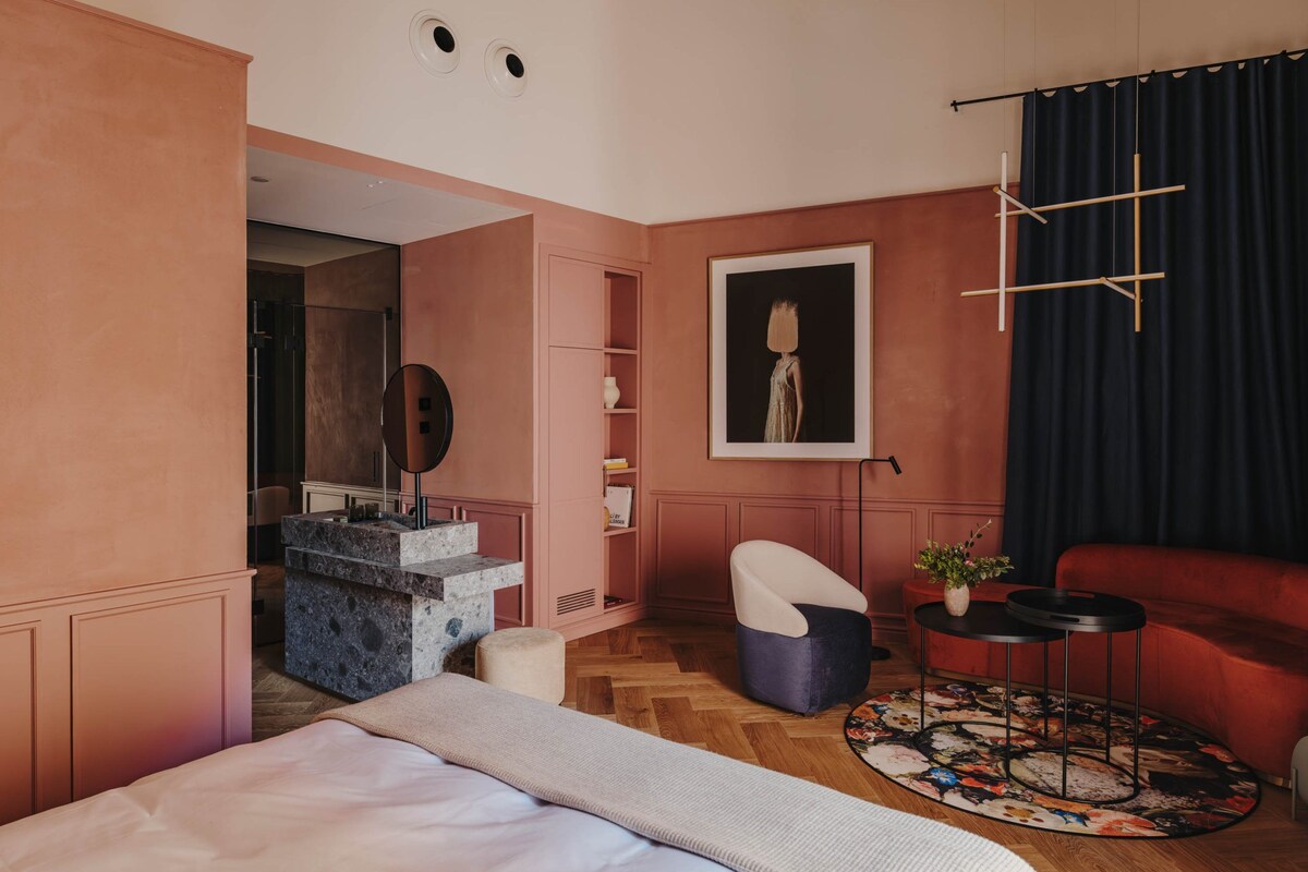 A modern bedroom with a bed, a vanity with a mirror, a red sofa, a round coffee table, and a painting on the wall.
