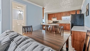 Un comedor con mesa y sillas de madera, una cocina con muebles de madera y un refrigerador, y una puerta que da a un baño.