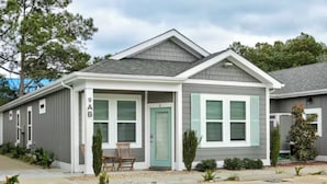 A two-story building with a grey exterior and a green door. It has a covered porch with two chairs and a small table.
