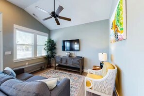 Un salon avec un ventilateur de plafond, un téléviseur, un canapé, un fauteuil, une plante et une fenêtre avec des stores.