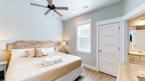 A bedroom with a bed, a ceiling fan, a nightstand, a lamp, a window with blinds, and a door. - The Great Crabsby | Dogs OK! Walk to beach, Pool (Corolla)