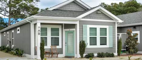A two-story building with a grey exterior and a green door. It has a covered porch with two chairs and a small table.